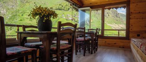 A wooden interior with a table and chairs, a vase with flowers, and a view of a green valley through large windows.