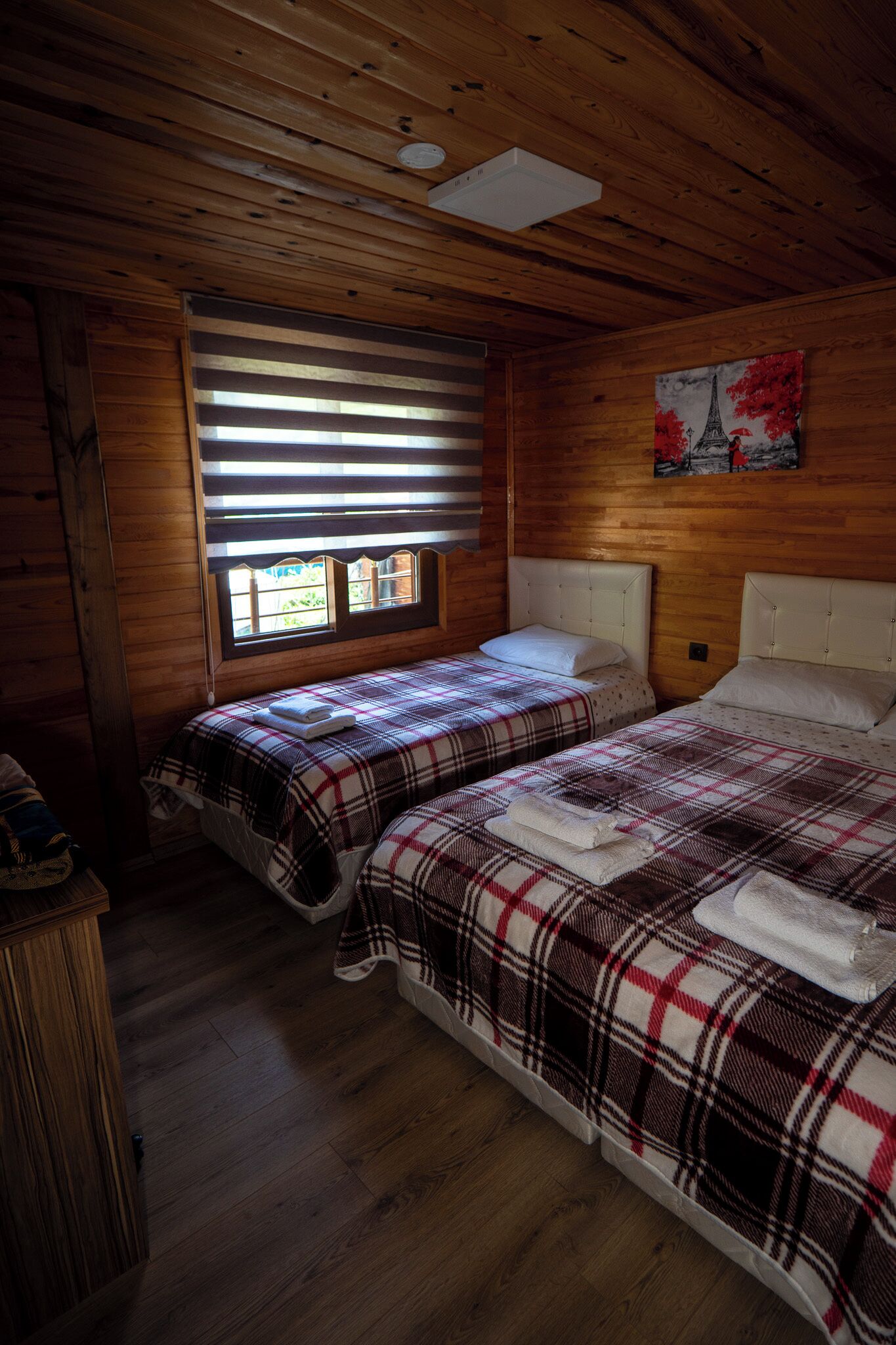 Une chambre de style cabane avec deux lits, des murs en bois, une fenêtre avec des stores et un tableau représentant la Tour Eiffel.