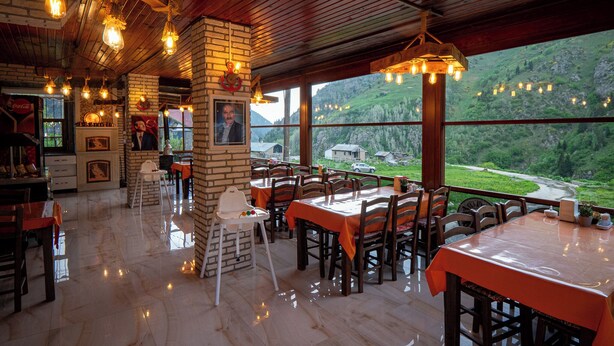 Un restaurante con techo de madera y paredes de ladrillo, mesas con manteles anaranjados y vistas a un valle verde.