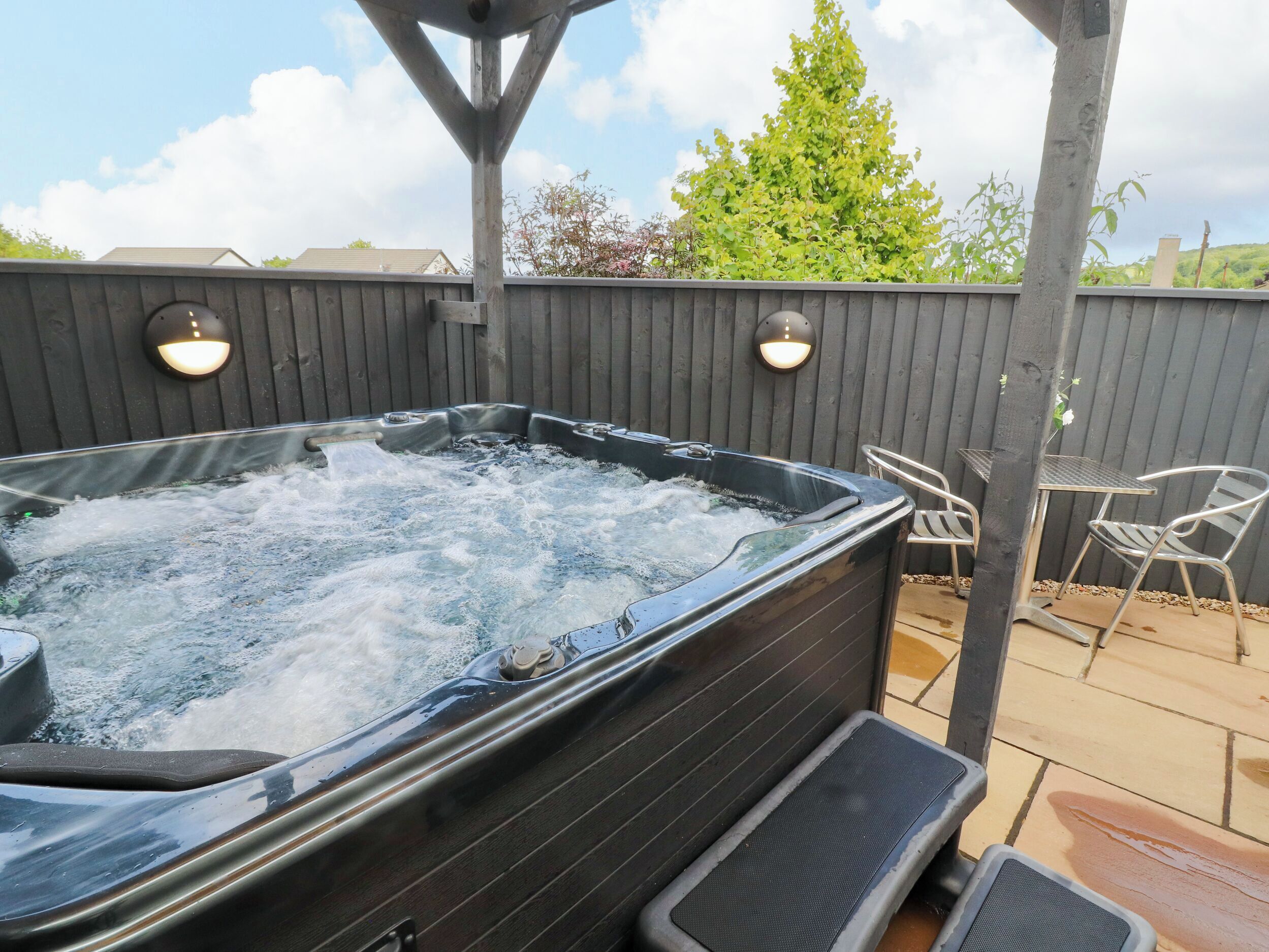 Un jacuzzi con cubierta de madera, dos luces fijadas a la pared y una mesa de patio con sillas.