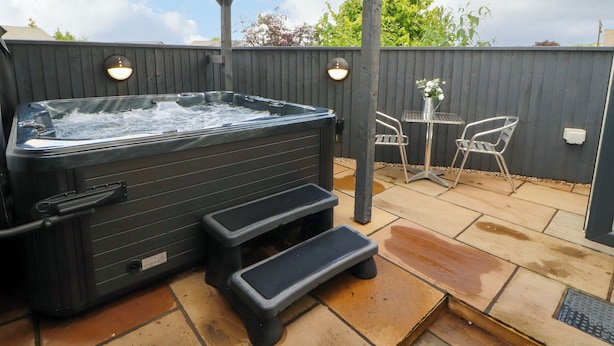 Un jacuzzi bajo una pérgola de madera con dos escalones de acceso, una mesa con sillas y una valla de madera al fondo.