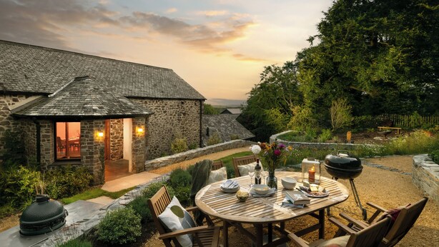 A stone house with a patio area featuring a round wooden table set for dining, surrounded by chairs.