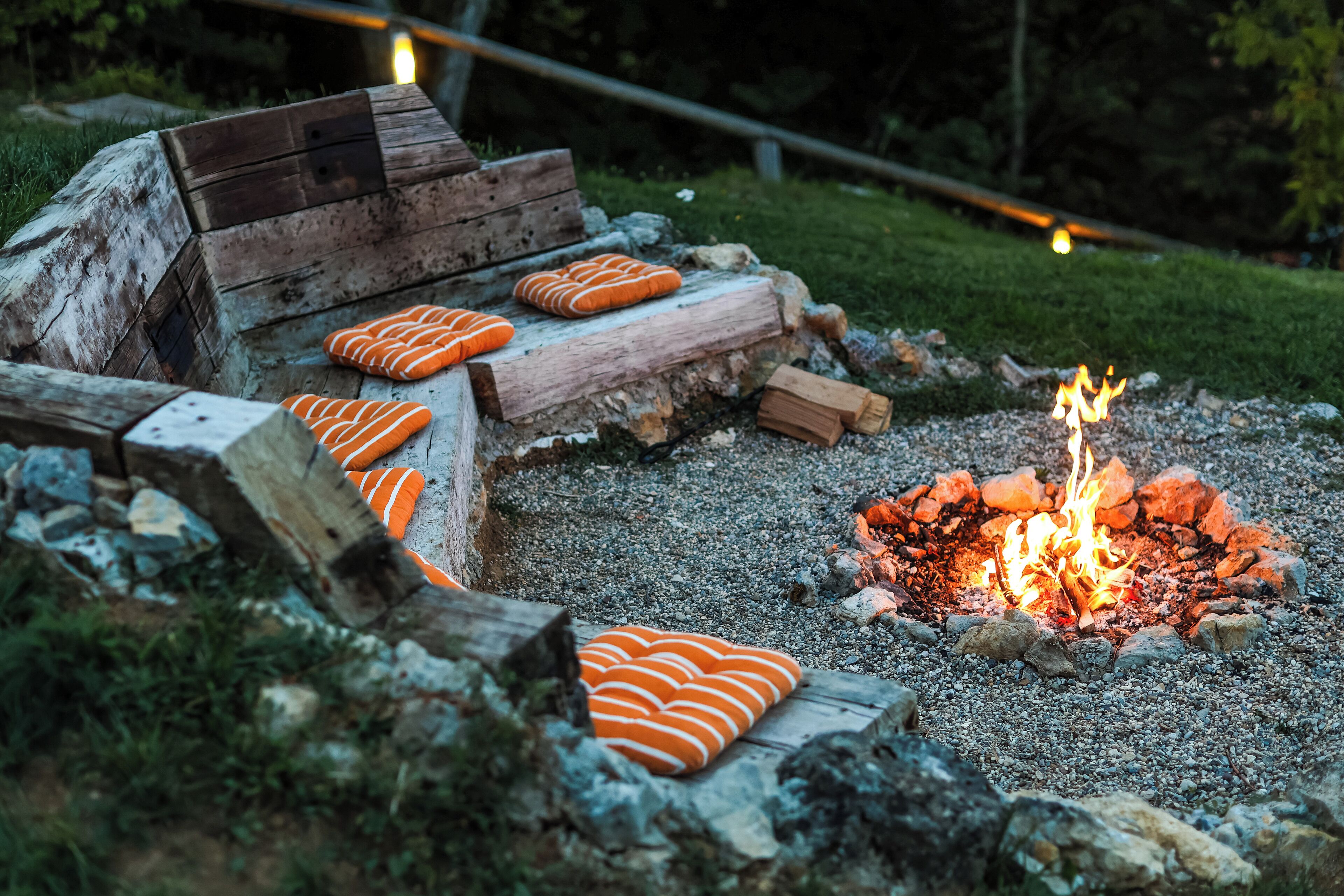 A rustic outdoor seating area with a fire pit, surrounded by logs and orange cushions.