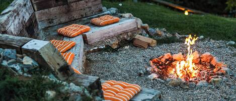 A rustic outdoor seating area with a fire pit, surrounded by logs and orange cushions.