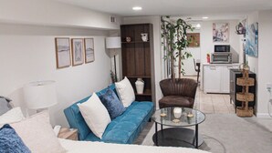 Een moderne woonkamer met een groot bed, een blauwe bank, een glazen salontafel, een bruine leren fauteuil en een keuken in de achtergrond.
