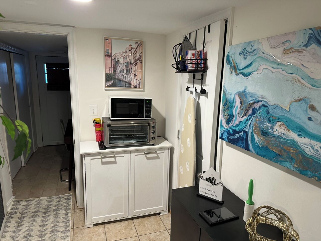 A compact kitchen with a microwave, toaster, and a small cabinet. There is a painting on the wall and a Wi-Fi sign on the counter.