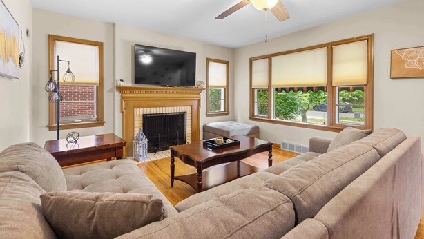 A living room with a fireplace, a television, a ceiling fan, and a large window with a view of greenery. - Your friendly neighborhood Airbnb (Rockford)