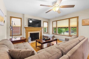 Ein Wohnzimmer mit Kamin, Fernseher, Deckenventilator und einem großen Fenster mit Blick auf Grünflächen.
