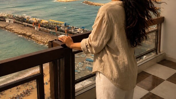 Eine Person steht an einem Fenster mit Blick auf einen Strand, einen Steg und einen wolkenlosen Himmel. Im Raum ist ein Schachbrettfußboden.