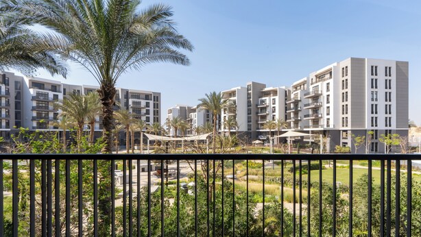 A modern residential complex with multiple apartment buildings, a palm tree, and a fenced garden area.