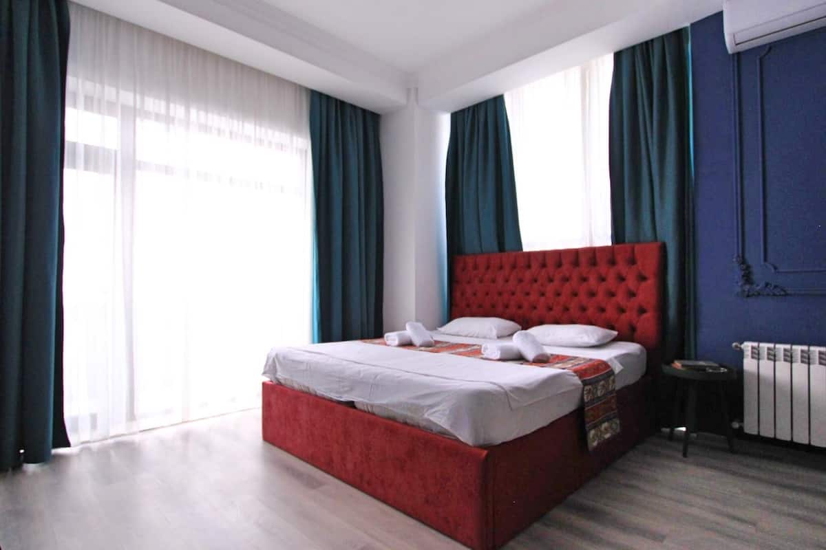 A modern hotel room with a red bed, white linens, and a view of the city through large windows with blue curtains.