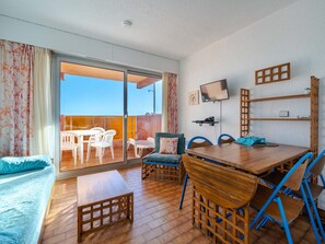 A room with a wooden floor, a sofa, a dining table with chairs, a TV mounted on the wall, and a balcony with a view.