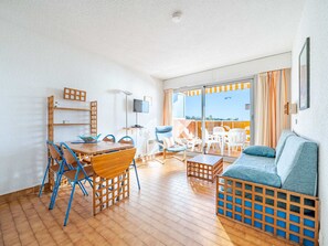 Un vaste espace de vie comprenant une table à manger en bois, des chaises bleues, un canapé bleu et un balcon agrémenté de chaises blanches et de rideaux orange.