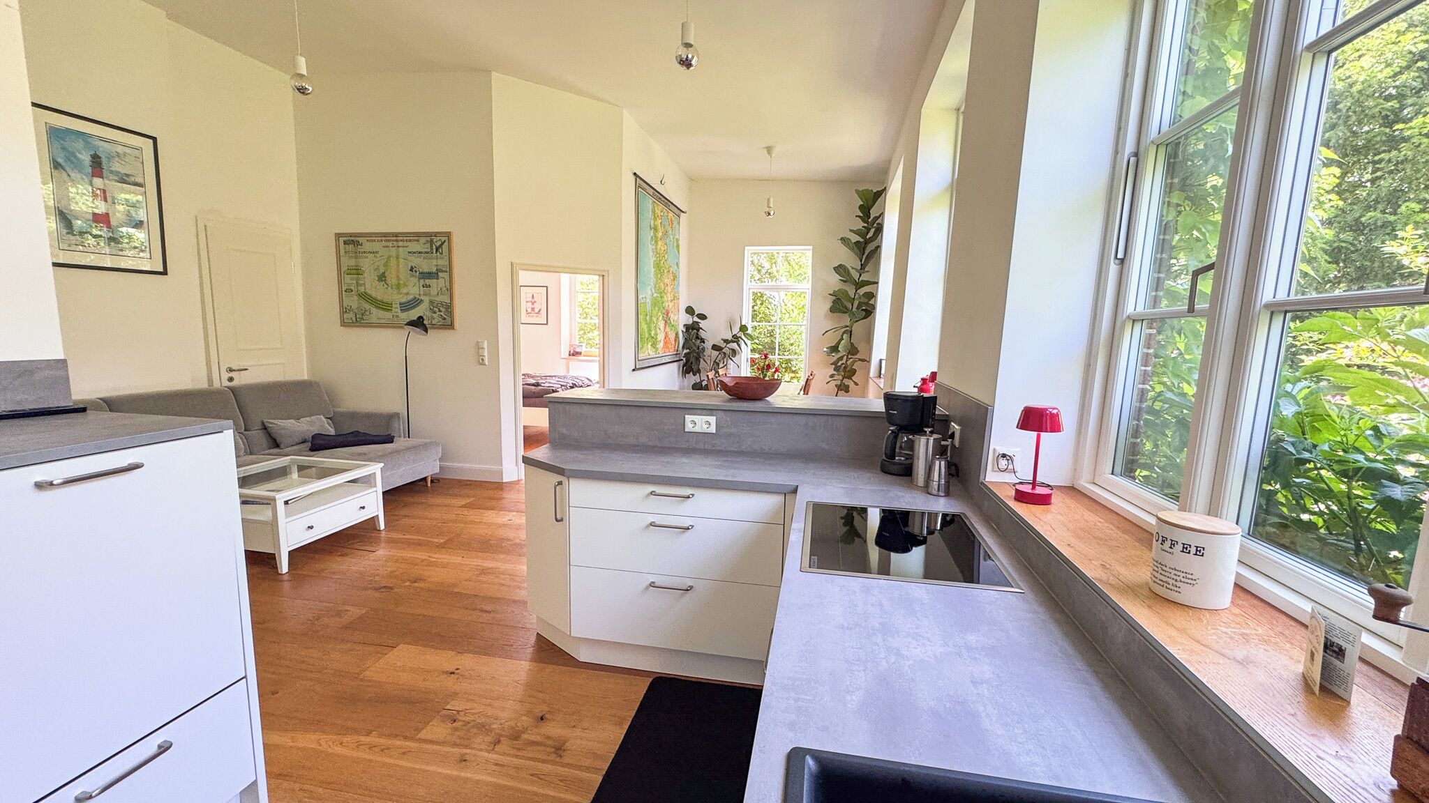 A modern kitchen with a dining area, wooden flooring, and large windows offering a view of greenery.