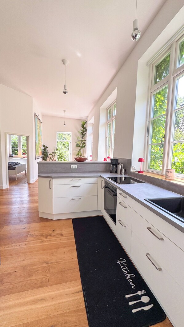 A modern kitchen with white cabinets, a black countertop, and a black rug with 'Kitchen' written on it. There are windows with greenery outside, and a coffee maker on the counter.