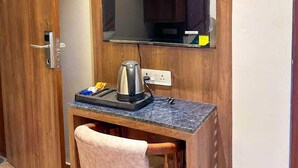 Habitación compacta con un armario de madera, un escritorio pequeño con hervidor, una silla y televisión montada.