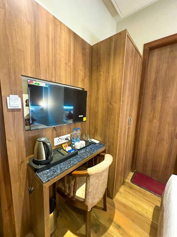 A compact hotel room with a wooden wardrobe, a small desk with a kettle, a television, and a chair.