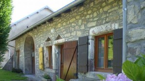 Un edificio de piedra con varios arcos en puertas y ventanas, un pequeño jardín con una planta en maceta y una flor rosa al frente.