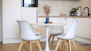 Una cocina moderna con armarios blancos, una mesa redonda blanca con cuatro sillas blancas, un jarrón azul con flores secas y una cafetera sobre la encimera.
