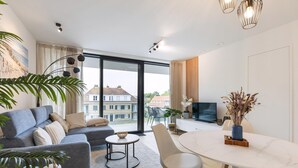 Una sala de estar moderna con un sofá, una mesa de comedor y un televisor. Hay plantas y un ventanal con vistas a edificios.