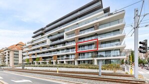 Un edificio de varios pisos con balcones, un paseo peatonal y una vía ferroviaria al frente.