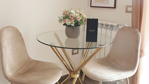 Et spiseområde med et glasbord, to stole, en vase med blomster og indrammede kunstværker på væggen.