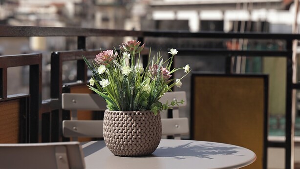 Et rundt bord med en flettet kurv med blomster, en stol og et bybillede i baggrunden.