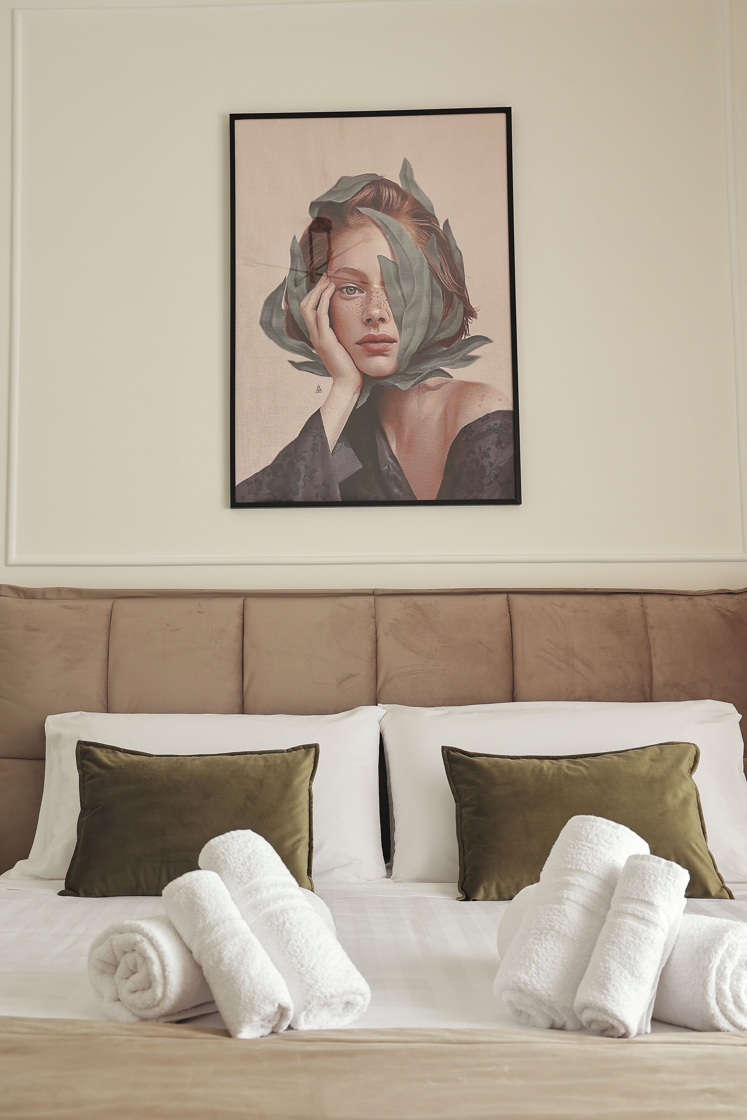 Una cama bien hecha con sábanas blancas y dos almohadas verdes, una obra enmarcada sobre la cama, y dos toallas blancas enrolladas sobre la misma.