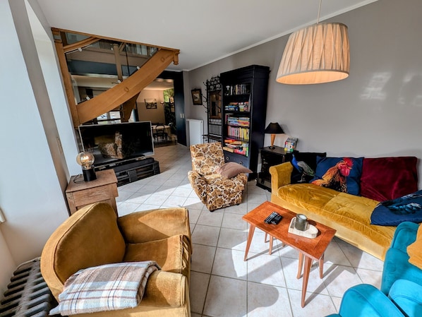 Un salon avec un canapé, un fauteuil et une table basse. On aperçoit une bibliothèque et un téléviseur au fond de la pièce.