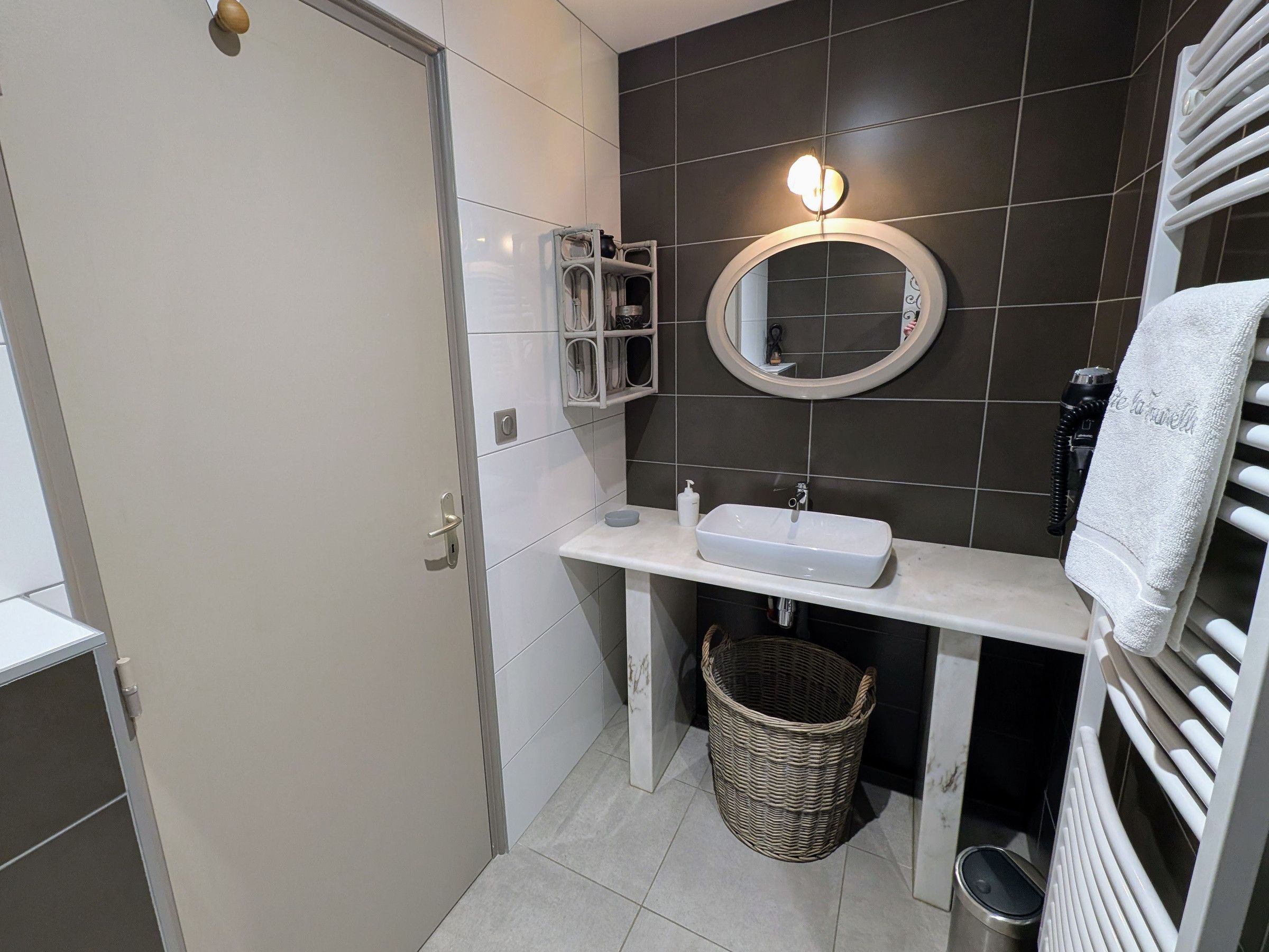 A bathroom with a white sink, dark tiled walls, a round mirror, a towel rack, and a wicker basket.