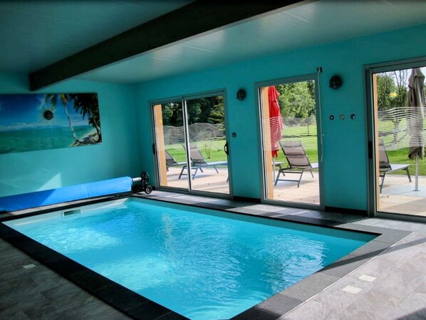 An indoor swimming pool with clear glass doors leading to an outdoor area with lounge chairs and a red umbrella. - Charming cottage in Ognon valley with spa (Bay)