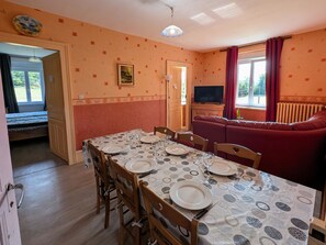 Ein Essbereich mit Tisch für vier Personen, Holzsesseln, einer Couch und einem Fernseher im Hintergrund.