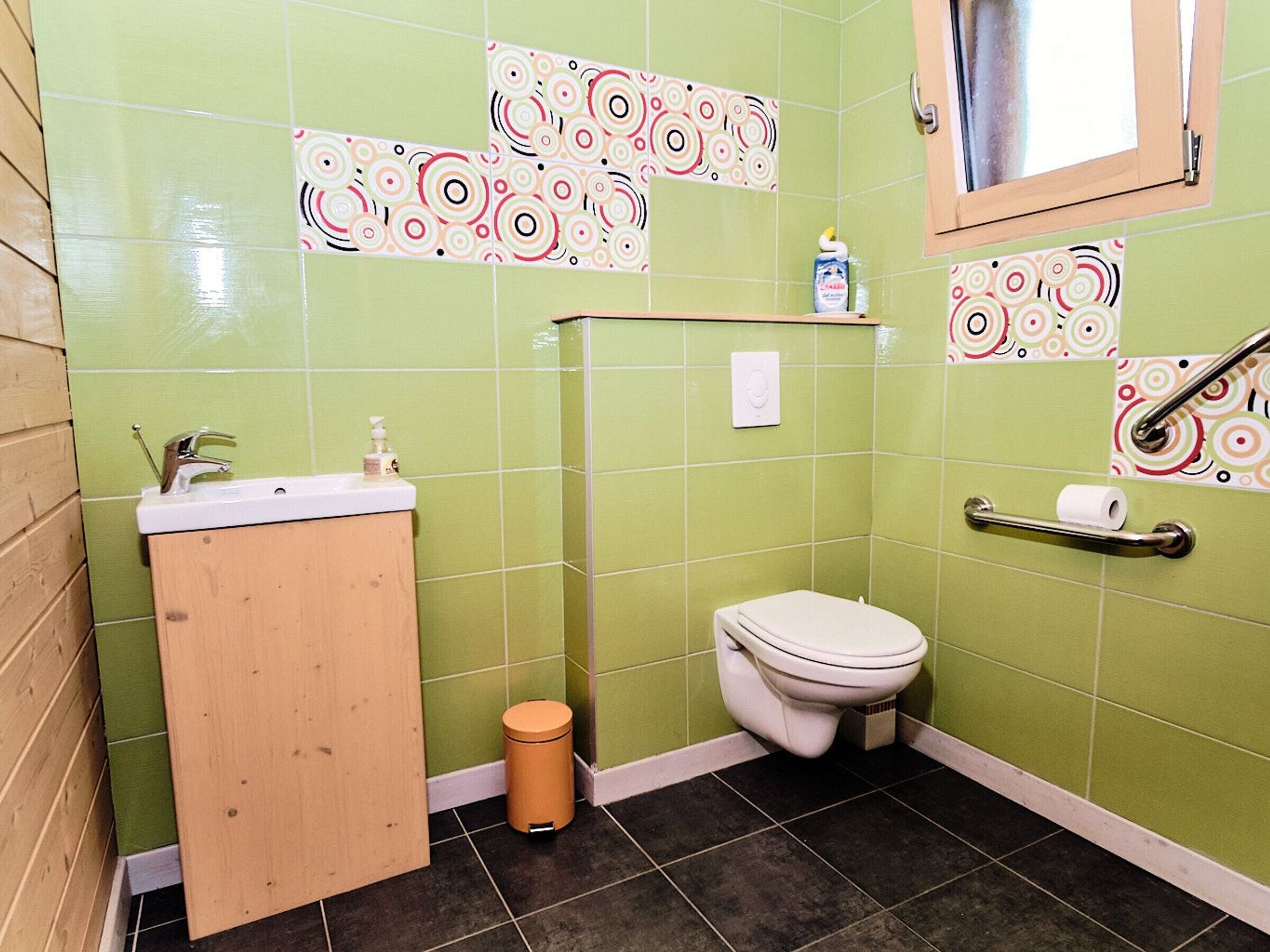 Une petite salle de bain avec des carreaux verts, un meuble en bois, des toilettes, une poubelle et une fenêtre.