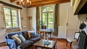Ein gemütliches Wohnzimmer mit einer Couch, einem Couchtisch und einem Kamin. Es gibt Fenster mit Vorhängen, eine Heizung und einen Kronleuchter.
