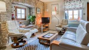 A living room with a stone wall, a fireplace, a television, and a large window with floral curtains. - Quiet cottage near Villersexel, nature and activities (Esprels)
