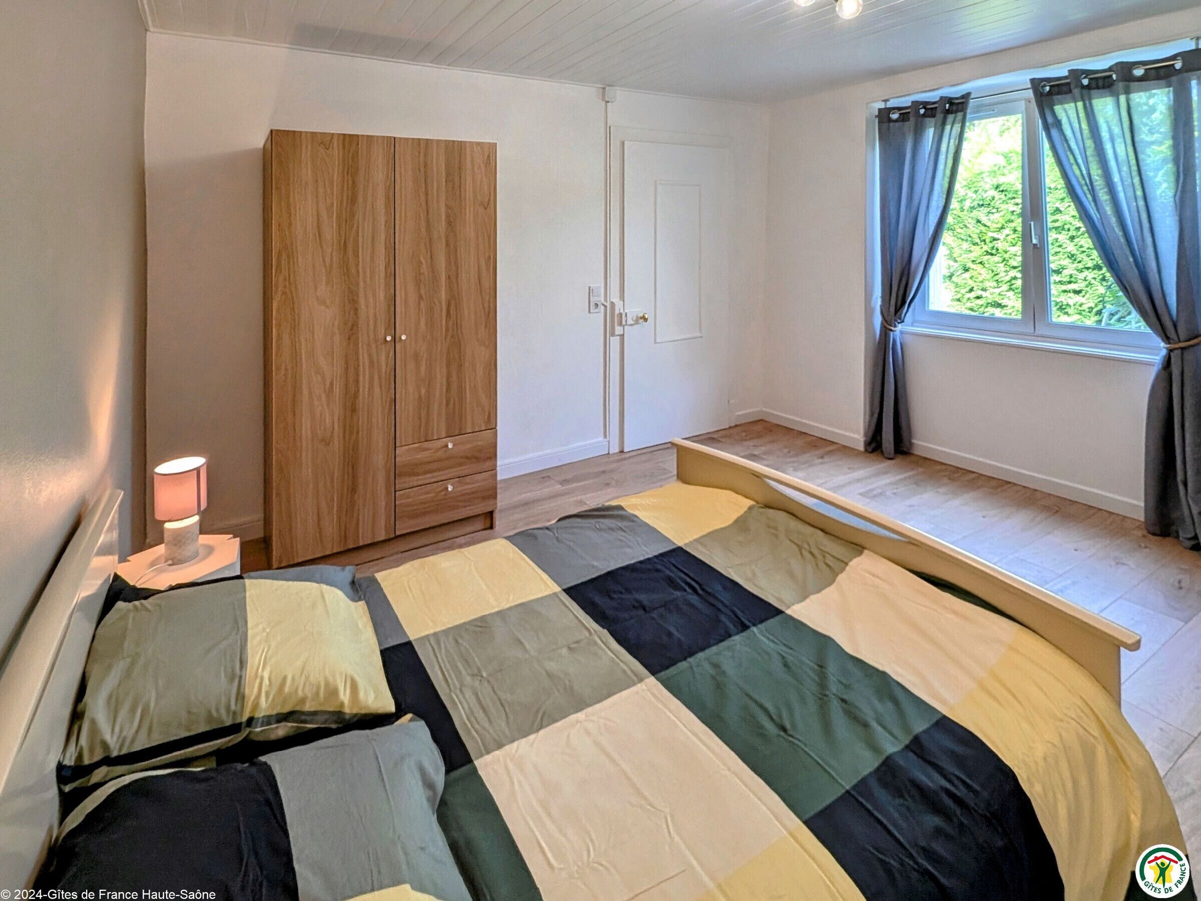 Un dormitorio con cama, armario, lámpara de noche y ventana con cortinas.