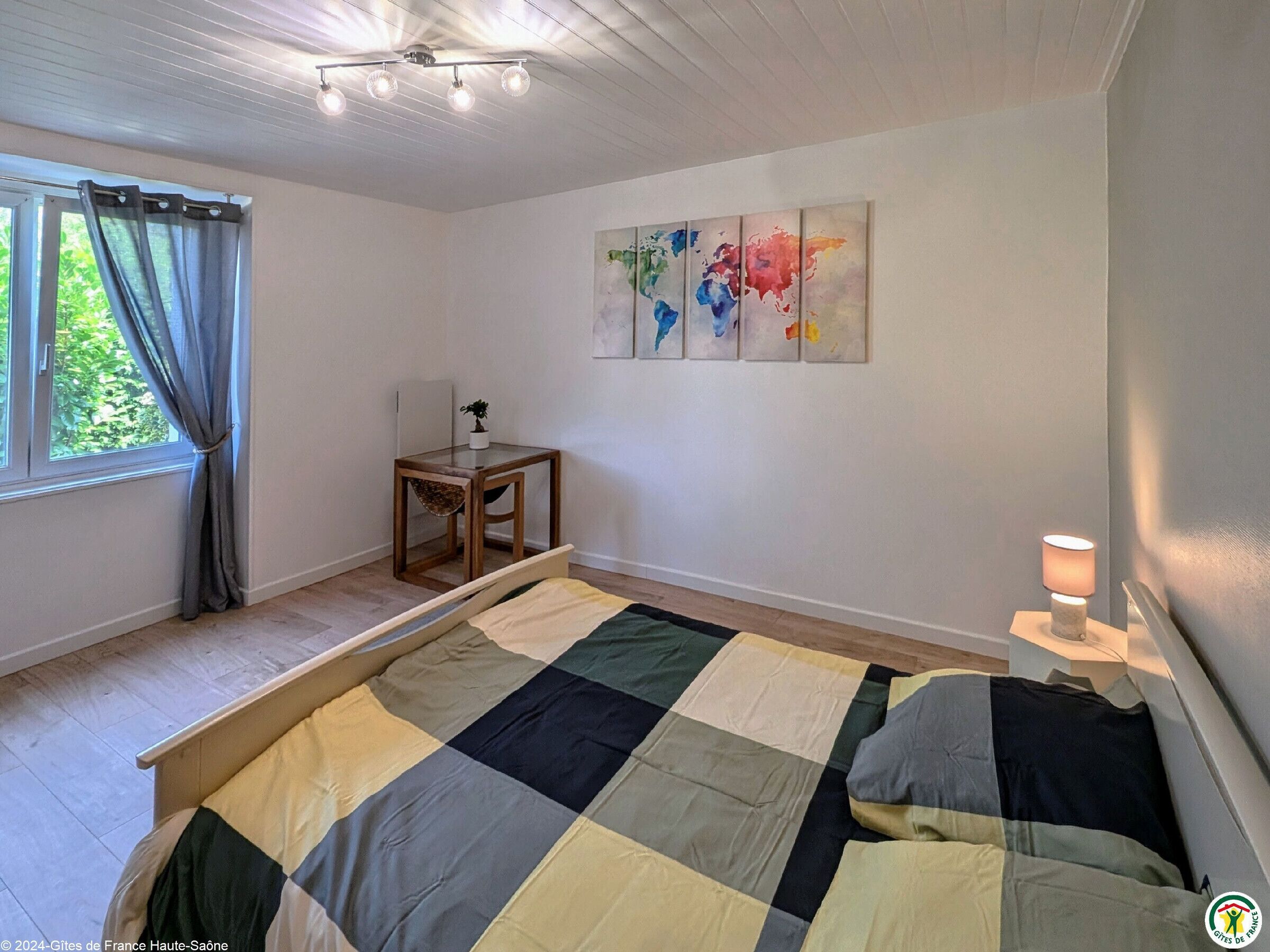 A bedroom with a large bed, a small table with a plant, a world map artwork, and a window with curtains.