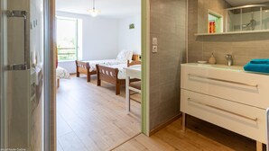 Une chambre moderne avec un plancher en bois, un lit recouvert d’une courtepointe blanche et colorée, une table de chevet et une salle de bain dotée d’une cabine de douche en verre et d’un meuble-lavabo blanc.
