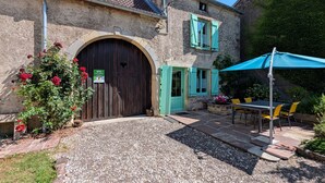 Un bâtiment rustique en pierre, doté d’une grande porte en bois, d’une petite fenêtre avec volets et d’un patio aménagé avec des sièges d’extérieur.