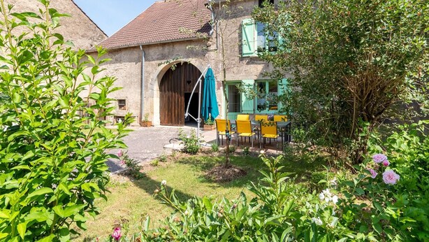 Un bâtiment rustique en pierre, doté d’une grande porte d’entrée en arc de cercle, d’un espace extérieur abrité avec des chaises jaunes, et d’un jardin où poussent diverses plantes et fleurs.
