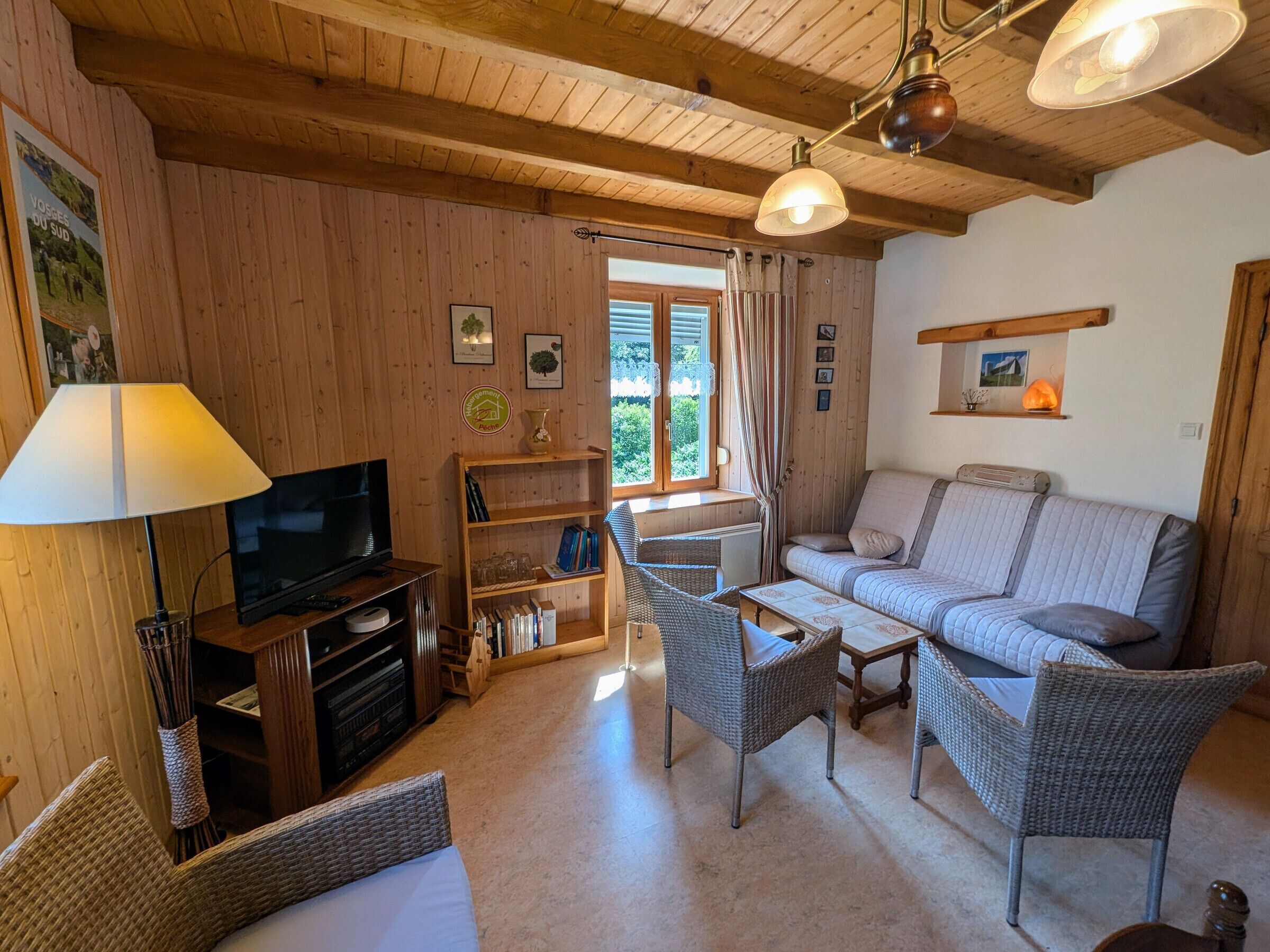 Een kamer met houten lambrisering, een bank, twee rieten fauteuils, een televisie, een boekenkast en een raam met gordijnen.