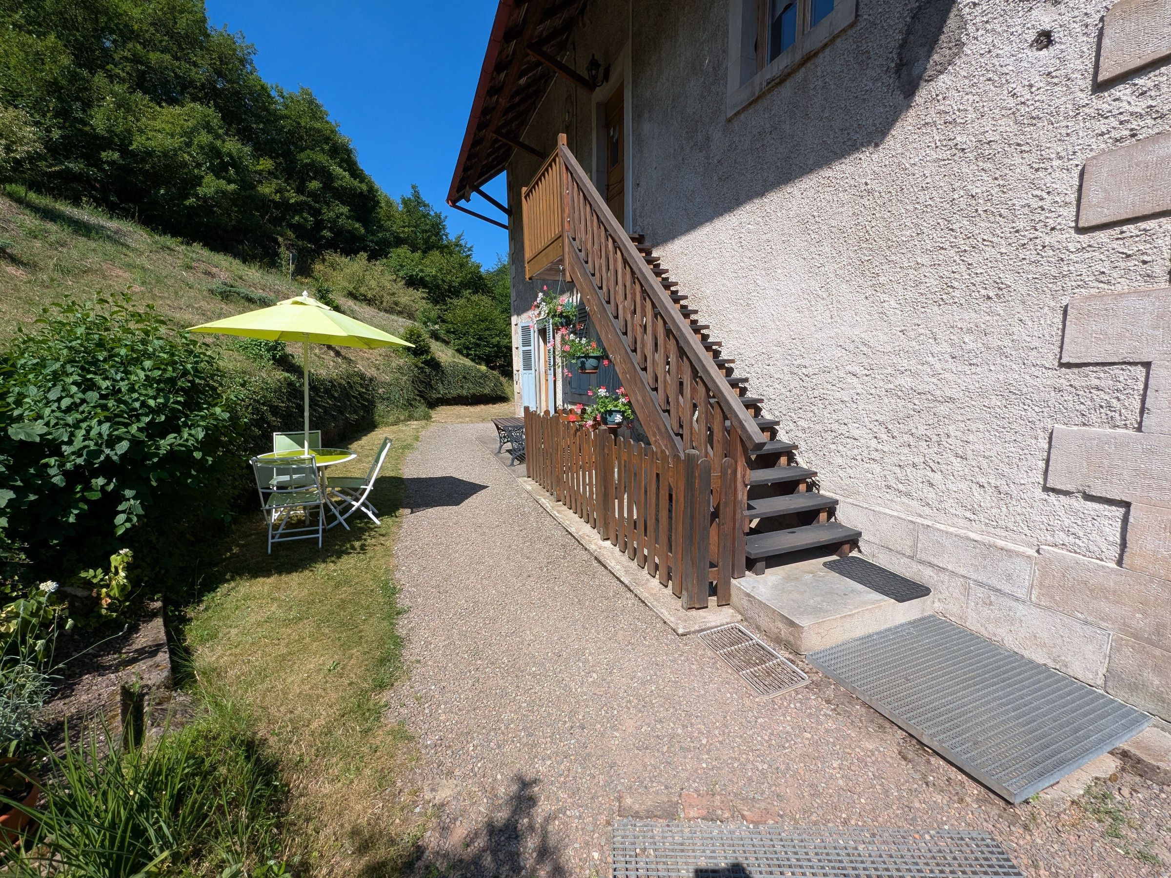 A wooden staircase leading to a building with a stone facade, a paved patio with a table and chairs, and a green umbrella.