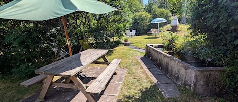 A garden with a wooden picnic table and benches, surrounded by lush greenery and a small water feature.