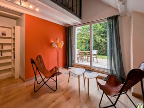 Un salon moderne doté d’une grande porte-fenêtre coulissante donnant sur une terrasse, d’une bibliothèque et de deux fauteuils en cuir.