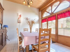 A dining area with a table set for four, wooden chairs, a vase with dried plants, a painting on the wall, and a window with red curtains. - Charming Cottage in the Vosges Saônoises (Saint-Bresson)