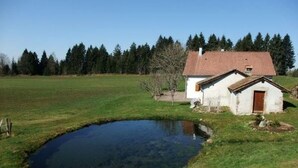 En lille hvid bygning med rødt tag, en sø og en skov i baggrunden.