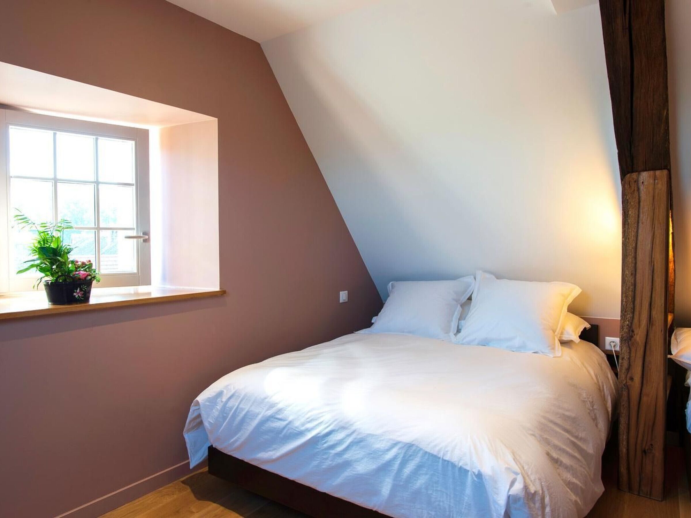 A bedroom with a sloped ceiling, a window with a potted plant, and a bed with white bedding.