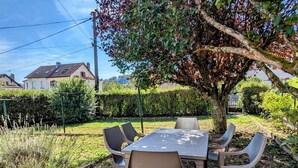 An outdoor dining area with a table and chairs under a tree, surrounded by a garden with various plants and a hedge.