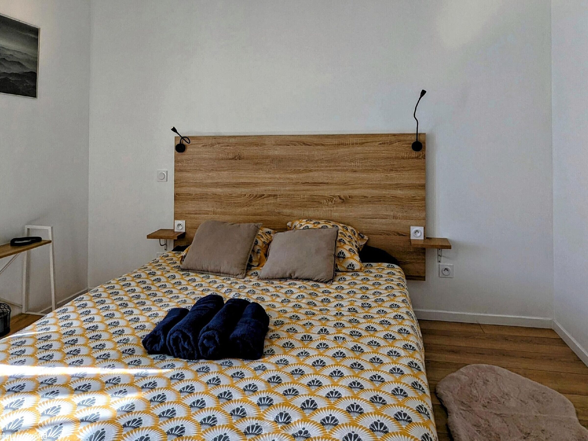 Un dormitorio con cabecera de madera, colcha estampada, dos almohadas y una toalla doblada sobre la cama.