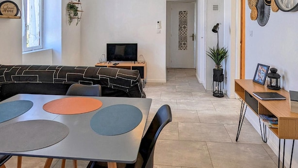 A modern living room with a sectional sofa, a dining table with colorful placemats, a TV on a wooden stand, and decorative wall art.